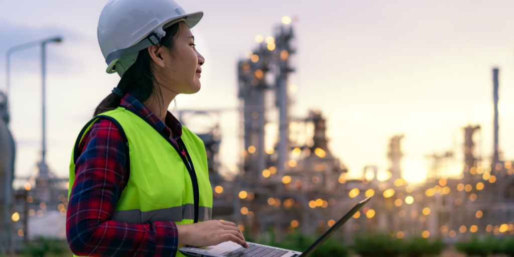 Female engineer in safety gear, working on laptop with refinery in the background