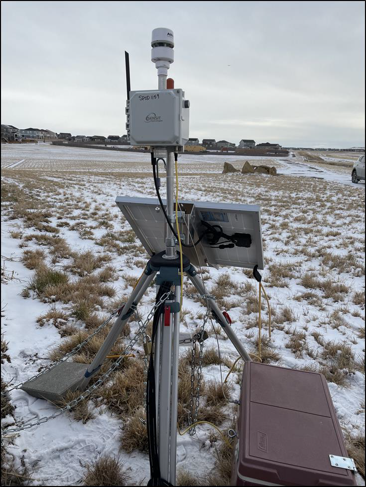 Sensor Pod mounted on a tripod in a field near a residential area, with a solar panel and battery as well as a meteorological station for wind speed and directions data.